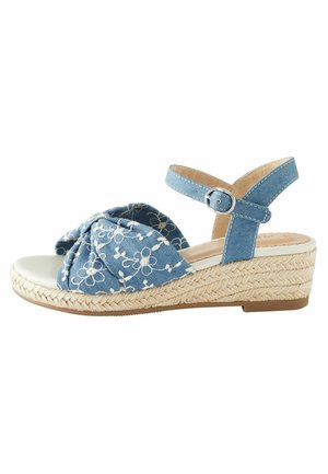 Sandale compensée avec dessus en tissu bleu, broderie florale blanche, bride à la cheville avec boucle, et semelle en jute tressée sur fond blanc.