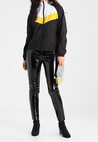 Černá větrovka s bílými a žlutými panely, zkombinovaná s lesklými černými legínami. Modelka drží stříbrný psaníčko. Černé kotníčkové boty s modrým podpatkem.