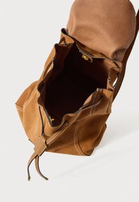 Hnědá semišová taška se stahovací šňůrkou, koženým popruhem, bílým prošitím a zlatým stiskacím uzávěrem, zobrazená otevřená z vrchu na bílém povrchu.