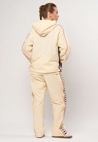 Kvinde iført cremehvid hættejakke og matchende joggingbukser med sorte og røde logostriber, stående med ryggen til.