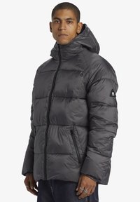 Vadderad grå jacka med huva, med dragkedjestängning och sidofickor. Slät, glänsande tyg med en strukturerad yta.