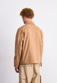 Beige denimjakke med strukturert krage og subtil stitching, med en avslappet passform og kneppede mansjetter, båret over cargo-bukser.