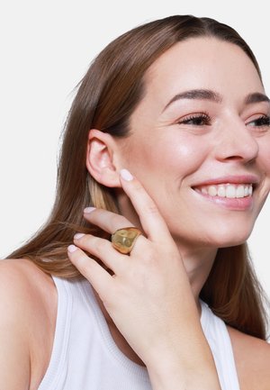 Anello color oro con forma organica, texture liscia e finitura lucida, indossato su una mano con unghie di colore chiaro e una canotta bianca.