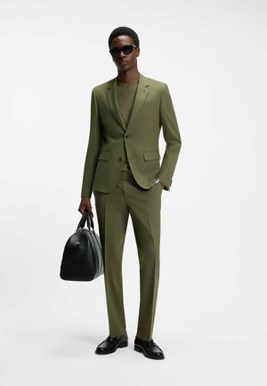 Man wearing olive green suit, matching shirt, black sunglasses, and black shoes, holding black leather bag in right hand.