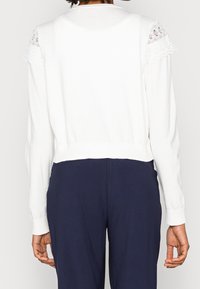 Bílý top s dlouhým rukávem s krajkovou lemovkou na švech ramen, žebrovaný lem a manžety; hladká textura; krátký střih. Kombinováno s námořnicky modrými kalhotami.