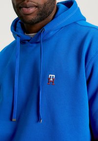 Felpa blu realizzata in morbido cotone. Presenta un cappuccio con lacci, tasca frontale e logo ricamato in rosso e bianco sul petto.