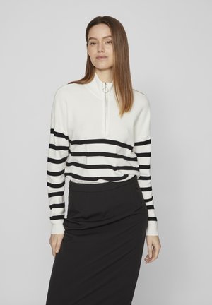 Sweater de rayas blancas y negras con cuello alto y cremallera frontal, combinado con una falda negra. Textura de punto suave y ajuste relajado.