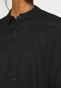 Chemise noire boutonnée avec un col montant, en tissu lisse. Présente un léger éclat et une fermeture classique à boutons assortis noirs.