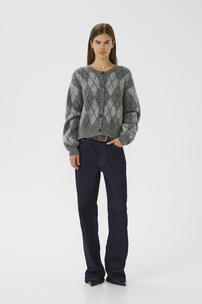 Kvinna klädd i en grå argyle-knäppt cardigan, mörkblå jeans och svarta spetsiga skor, står med en hand i fickan.