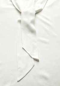 Weißes Blusentop mit einer Frontknotendetail, das eine weiche, glatte Textur und einen drapierten Schnitt aufweist. Der Stoff hat einen dezenten Glanz und ist leicht.