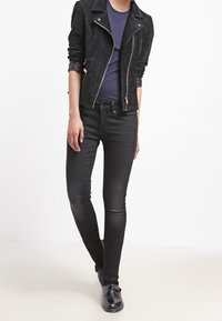 Chaqueta de moto de ante negra con herrajes en oro rosa, llevada sobre una blusa ajustada azul marino y pantalones skinny negros, combinada con zapatos negros.