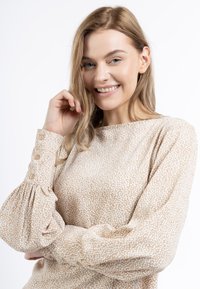 Beige lange mouwen top met een gestructureerd stippenpatroon en knoopdetails op de mouwen, met gathered stof bij de mouwen.