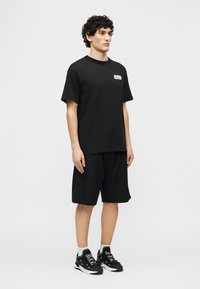 Νεαρός άνδρας που στέκεται, φορόντας μαύρο T-shirt με λογότυπο, μαύρη βερμούδα, λευκές κάλτσες και μαύρα αθλητικά παπούτσια με λευκά γράμματα στα κορδόνια.