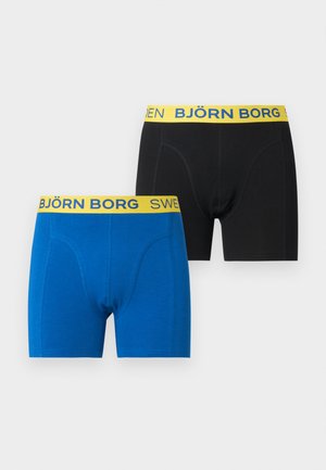 Λεπτός μυώδης μαύρος άνδρας που στέκεται ξυπόλητος, φοράει μαύρα μποξεράκια με κίτρινη μέση που γράφει "SWEDEN BJÖRN".