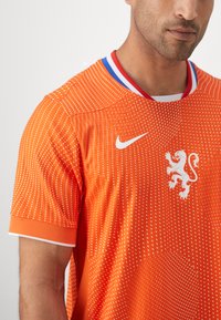 Nike voetbalshirt in feloranje met een gestippeld patroon, wit leeuwlogo en rood, wit, blauw gekleurde kraag en mouwaccenten.