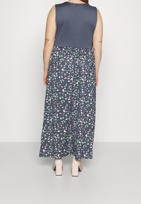 Maxi šaty s tmavošedým bezrukávovým topem a vzorovanou sukní, která má bílé a zelené abstraktní tvary na námořnickém pozadí.