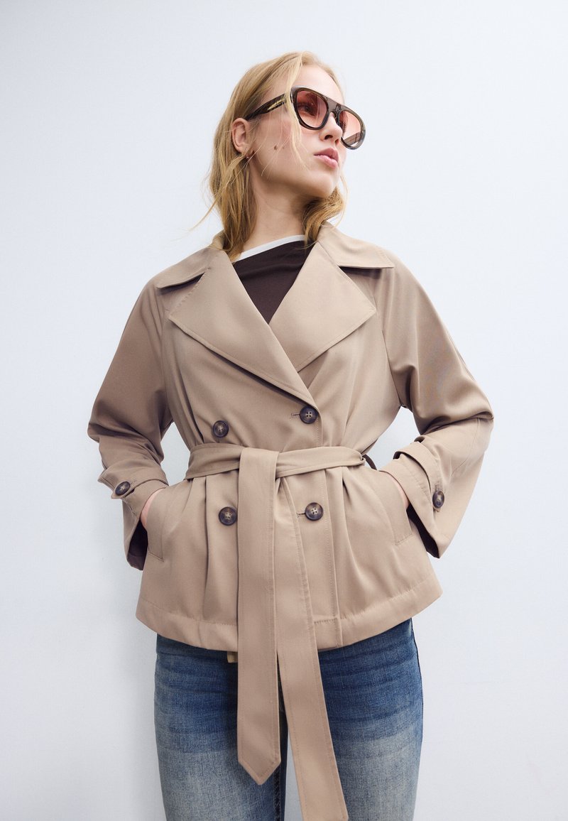 Femme portant un trench-coat beige ceinturé et des lunettes de soleil, posant avec les mains dans les poches sur un fond blanc uni.
