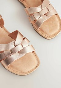 Sandalias metálicas de color rosa dorado con correas trenzadas, suelas planas y una textura de cuero suave, que cuentan con una abertura amplia para facilitar su uso.