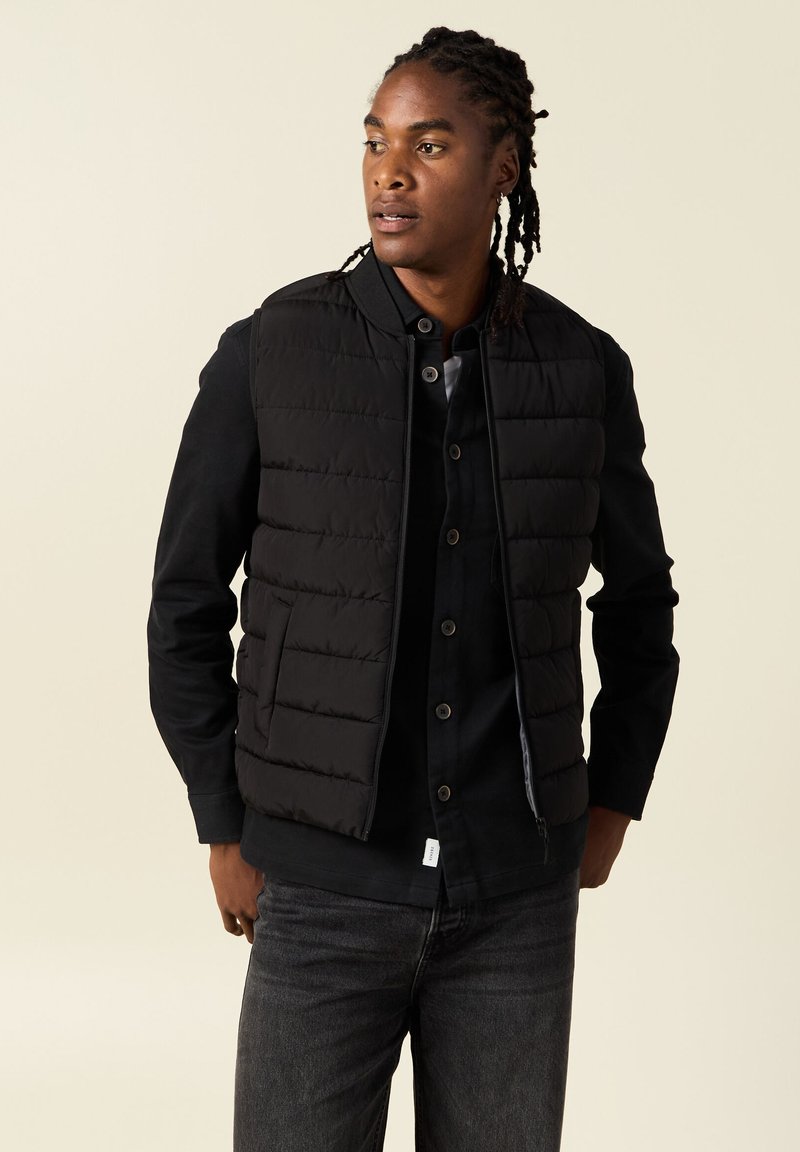 Chaleco acolchado negro con acolchado vertical, con cierre de cremallera y bolsillos laterales, usado sobre una camisa negra de botones y jeans oscuros.