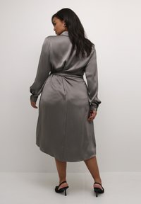 Graues Satin-Kleid mit langen Ärmeln und tailliertem Gürtel, mit einem Wickeldesign und subtiler Glanz, kombiniert mit schwarzen Sandalen mit Absatz.