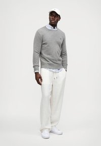 Suéter cinza de malha, camisa de vestir branca com padrão xadrez por baixo, calças de linho creme e ténis brancos. Boné branco na cabeça. Design simples.