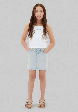 Chica con el pelo largo y castaño, lleva un top blanco de Calvin Klein, falda vaquera clara, sandalias beige, de pie con las manos en los bolsillos.