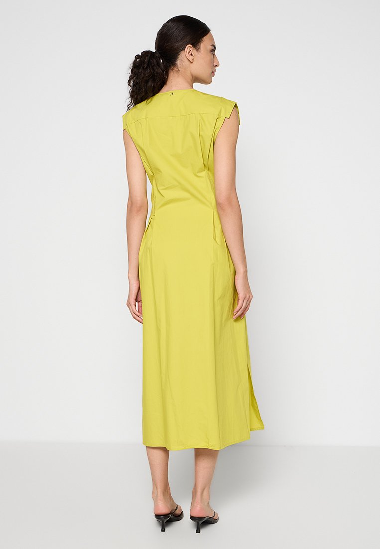 Kvinna med lockigt hår i en limegrön ärmlös midi-klänning och svarta högklackade sandaler, stående vänd bort mot en enfärgad bakgrund.