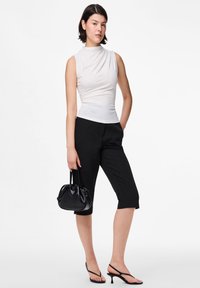 Biały top bez rękawów z marszczonymi detalami, zestawiony z czarnymi krótkimi spodniami. Modelka trzyma małą czarną torebkę i nosi czarne sandały na obcasie.