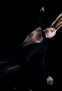Body nero con schiena trasparente, maniche lunghe e collo alto. I capelli fluiscono verso l'alto, su uno sfondo scuro. Pose dinamica e aerea.