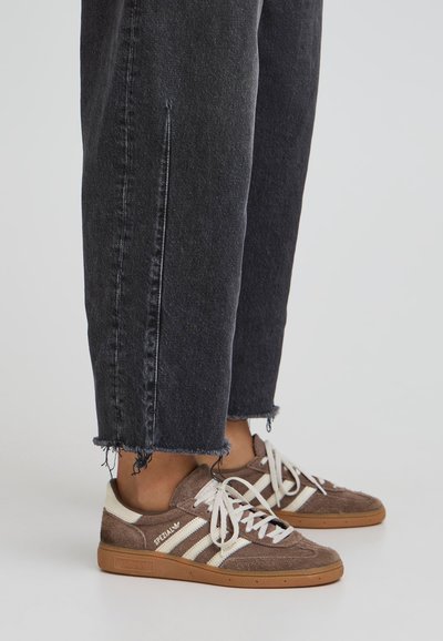 Rjavi semiš športni čevlji z belimi črtami, vezalkami in podplatom iz gumijastega materiala, v kombinaciji s temnimi jeans hlačami z raztrganimi robovi in stranskimi razpoke.