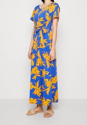 Vrouw draagt een blauwe maxi-jurk met grote gele bloemenprints, korte mouwen, een strikceintuur aan de voorkant en beige gevlochten instapsandalen.