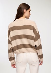 Sweter o rozluźnionym kroju w paski w odcieniach brązu i beżu, z obniżonymi ramionami i ściągaczami, zestawiony z jasnymi spodniami.