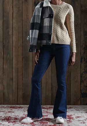 Person trägt cremefarbenen Zopfpullover, dunkelblaue ausgestellte Jeans, weiße Sneaker und hält eine schwarz-weiß karierte Jacke über der Schulter.