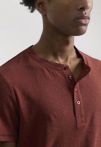 Camisa henley burdeos hecha de una tela suave, con cuello redondo, dos botones y mangas cortas con una textura suave.