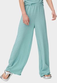 Licht turquoise wijde broek met een gestructureerd, golvend patroon, elastische tailleband en gegolfde zoom. Gepaard met zilveren slippers.