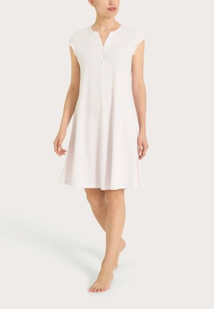 Donna che indossa un abito bianco a maniche corte, lungo fino al ginocchio, con bottoni sul davanti, in piedi a piedi nudi su uno sfondo neutro.