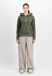 Olivno zeleni hudik z kenguru žepom in vrvicami, v kombinaciji s svetlo bež širokimi dihajočimi hlačami s stranskimi žepi in sproščenim krojem.