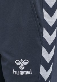 Marina sportbyxor med en texturerad väv, vitt chevronmönster längs sidan och en vit broderad "hummel"-logotyp.