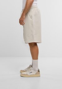 Bežové bavlnené šortky s elastickým pásom, bočnými vreckami a vyšívaným logom, kombinované s bežovými teniskami a svetlosivými ponožkami.