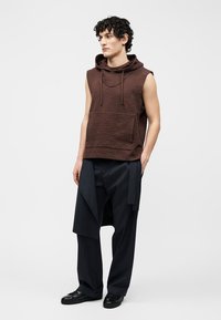 Jonge man met krullend haar, gekleed in een bruine mouwloze hoodie en zwarte broek met wijde pijpen en gelaagde stofpanelen, staand met handen in de zakken.