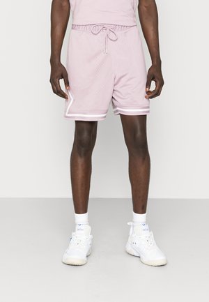 Shorts de sport en mesh rose avec une taille élastique, un détail de cordon de serrage et des accents de rayures blanches à l'ourlet. Portés avec des baskets blanches.