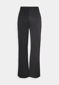 Pantalon noir avec un devant plat et une ceinture élastique, un côté uni noir, l'autre avec des rayures verticales blanches. Coupe ajustée à jambes larges.