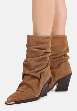 Bottes en daim marron décontractées mi-mollet avec des bouts pointus et des talons épais noirs portées sur des jambes nues.