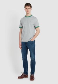 T-Shirt grigio a maniche corte con bordi verdi, abbinato a jeans blu e scarpe stringate marroni, in piedi contro uno sfondo neutro.