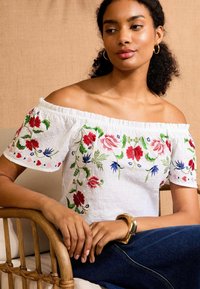 White off-the-shoulder top featuring intricate floral embroidery in red, blue, and green, with a textured fabric and elastic neckline.