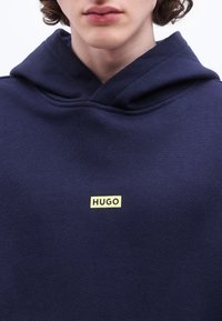 Νεαρό άτομο που φοράει μπλε σκούρο φούτερ με κουκούλα με το κίτρινο λογότυπο "HUGO" στο κέντρο του στήθους, μπροστά σε απλό λευκό φόντο.