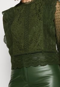 Top verde in pizzo con motivi floreali, dettagli testurizzati e bordi a scallop. Abbinato a pantaloni fitted in ecopelle verde.