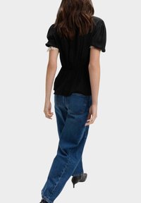 Schwarzes Oberteil mit Puffärmeln und tailliertem Schnitt, mit Spitzenbesatz; kombiniert mit lockeren blauen Jeans und schwarzen High-Heels.