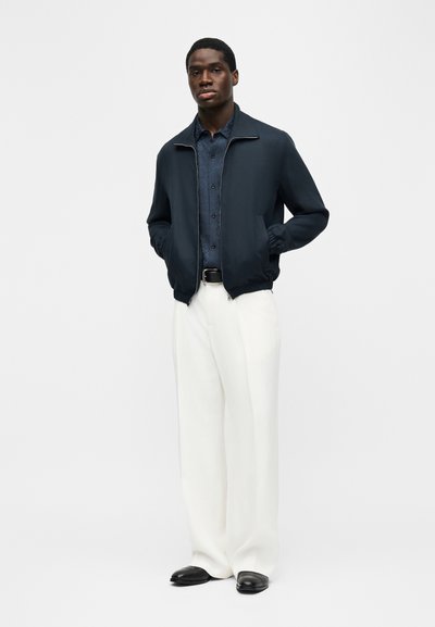 Homme portant une veste zippée bleu marine sur une chemise bleu foncé, un pantalon large blanc et des chaussures noires, debout les mains dans les poches.