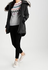 Svart parkas med huva kantad med päls, grå tröja med ett färgglatt diamantsmönster, svarta leggings och beige sneakers.